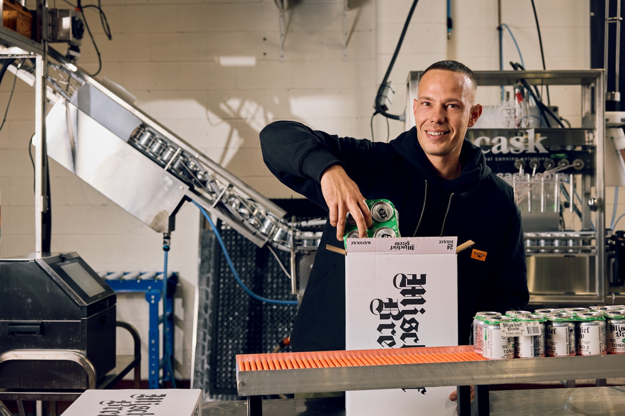A person by the Mischief brewing production line smiling to camera as they place cans into the carton